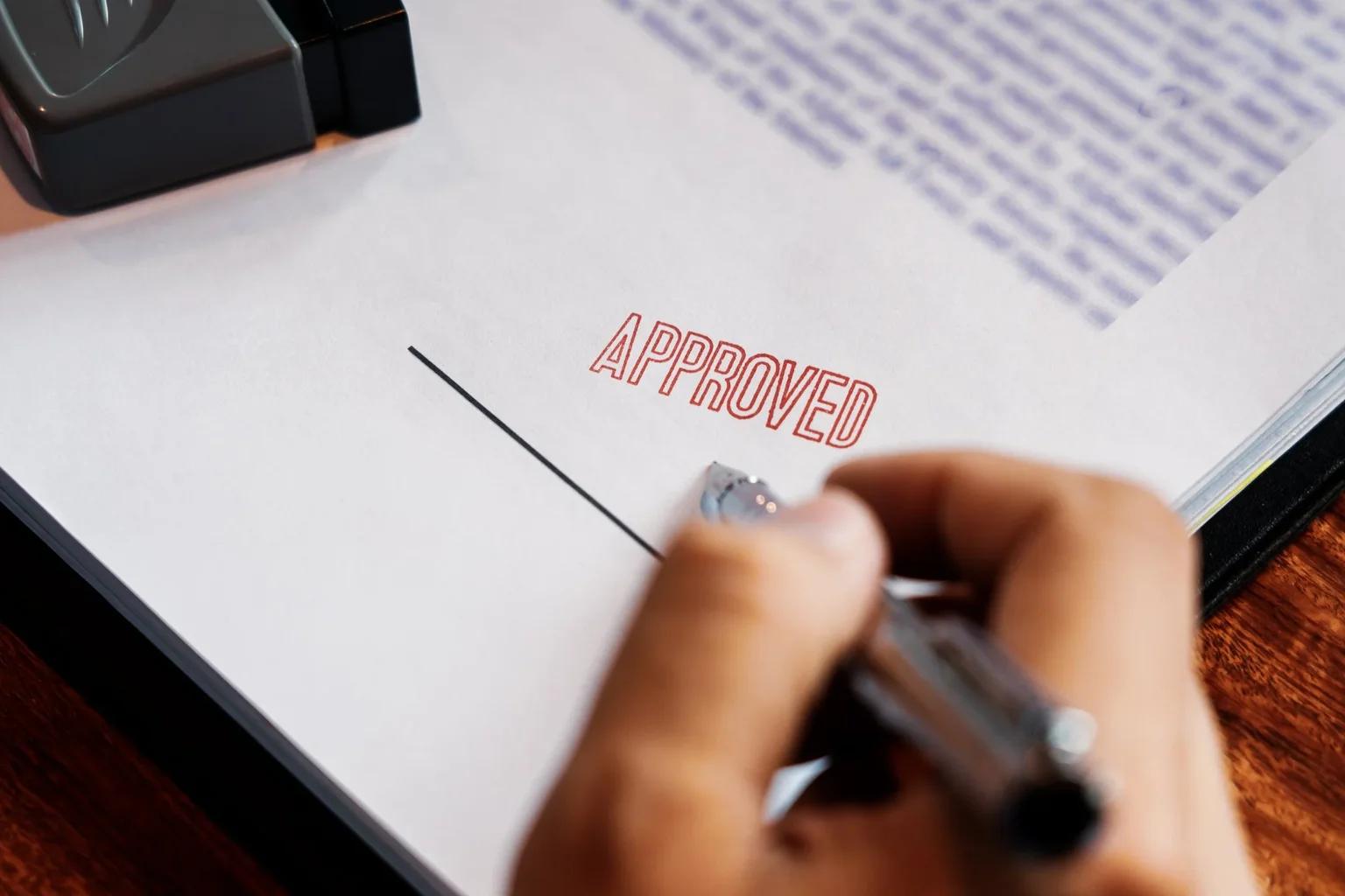 A close-up of a hand holding a pen signing an official document, with a black mechanical stamp and a prominent red APPROVED stamp, conveying a professional immigration approval.
